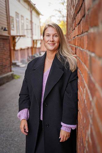 Linda Theologou in a black coat leaning against a brick wall smiling to the camera.