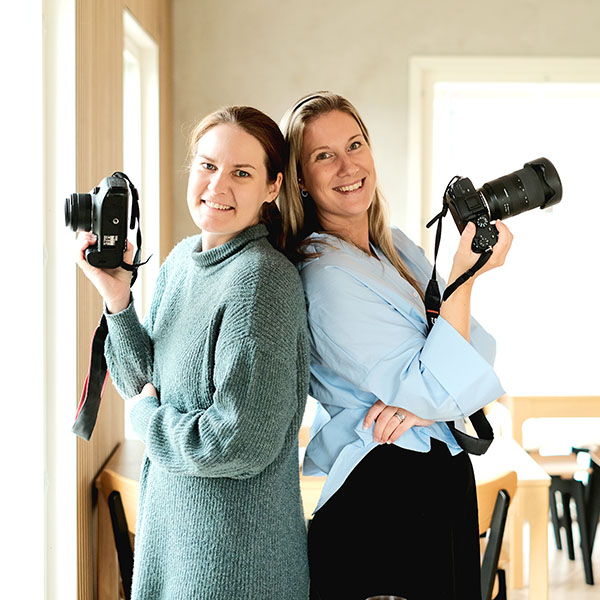 Linda and Satu holding cameras stand back-to-back in a well-lit room.