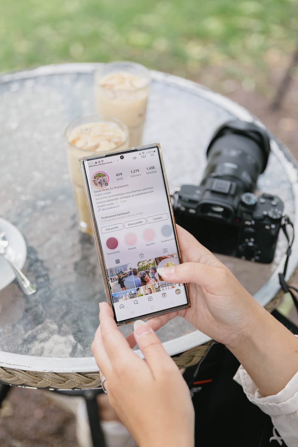 A person holds a smartphone displaying a social media profile at an outdoor table with iced coffee and a camera.