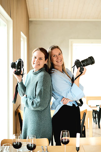 Linda and Satu back-to-back, each holding a camera. There are glasses of red wine on the table in front of them.